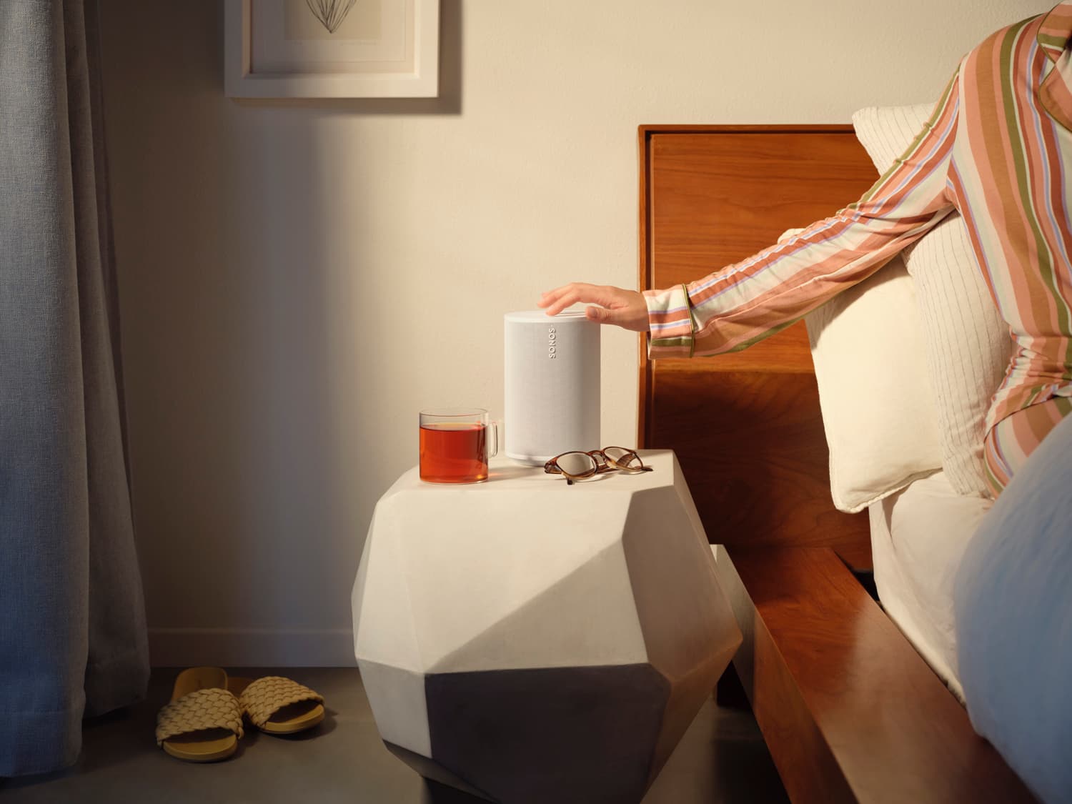 Hand reaching from bed to touch a white Sonos Era speaker on a geometric nightstand with tea and glasses
