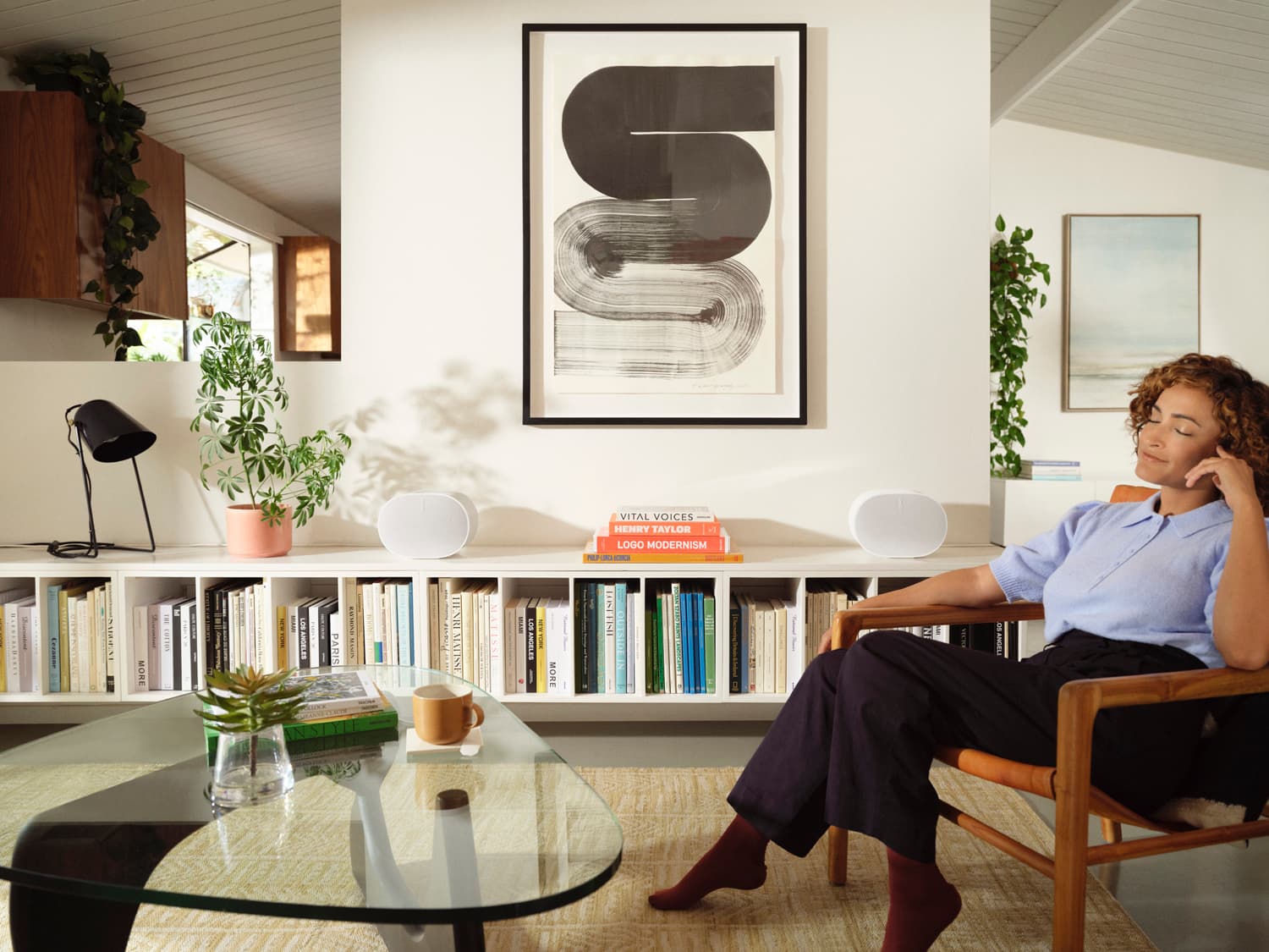 Woman relaxing in a chair in a mid-century living room with two white Sonos Era speakers on a bookshelf