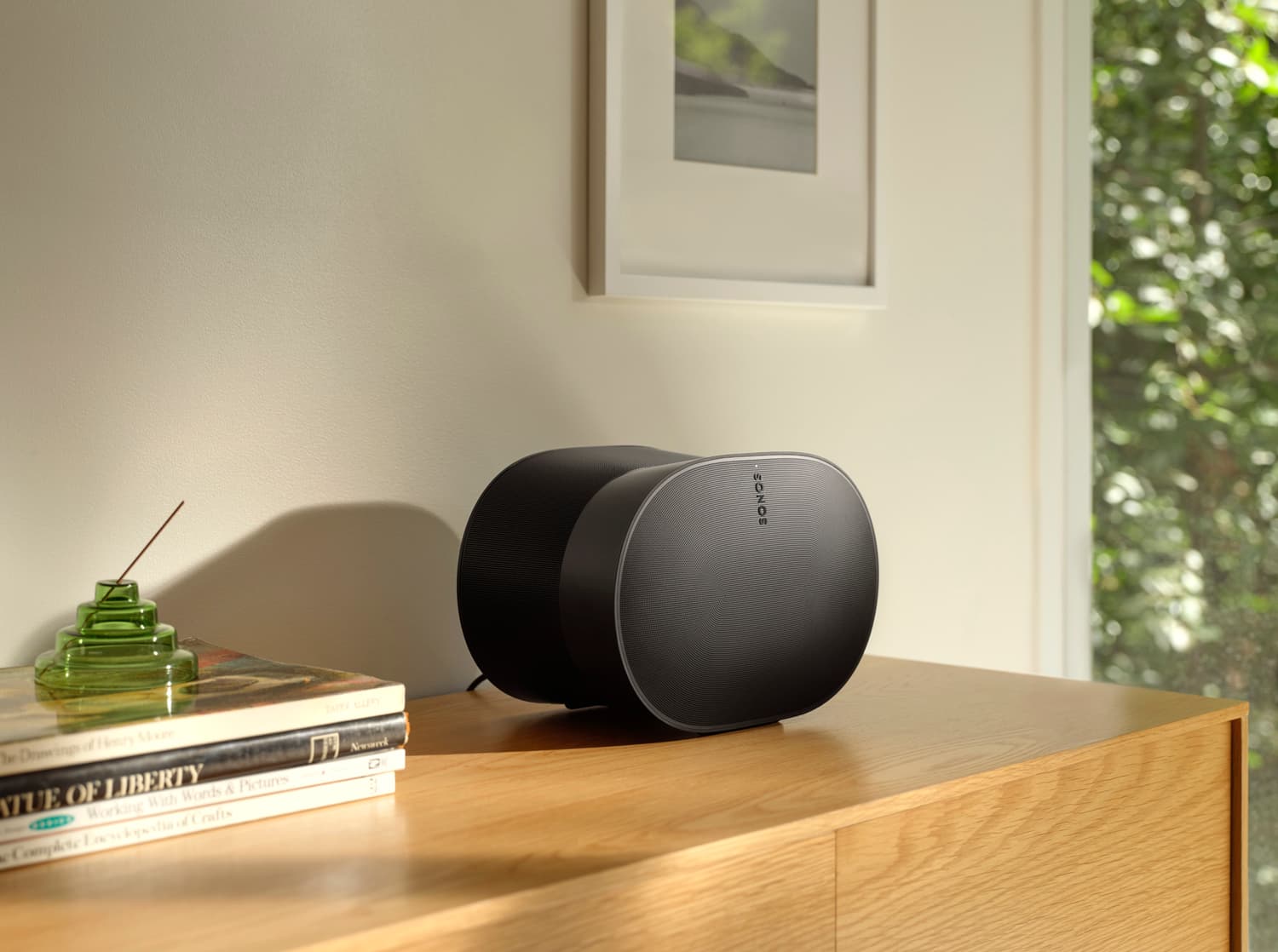 Black Sonos Era 300 speaker on an oak credenza beside stacked books and a green glass vase