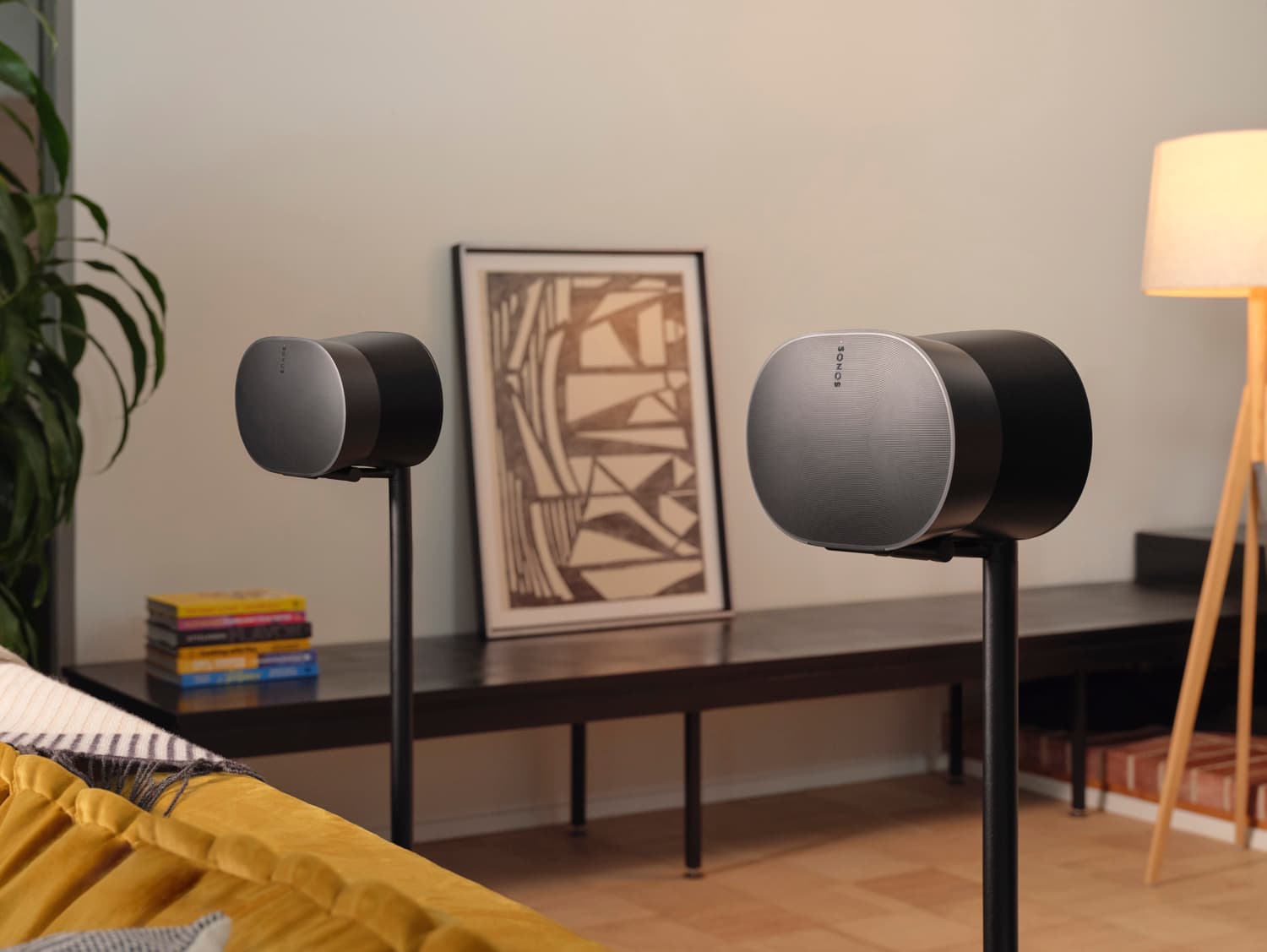 Two black Sonos Era 300 speakers on stands in a living room with framed art and a floor lamp